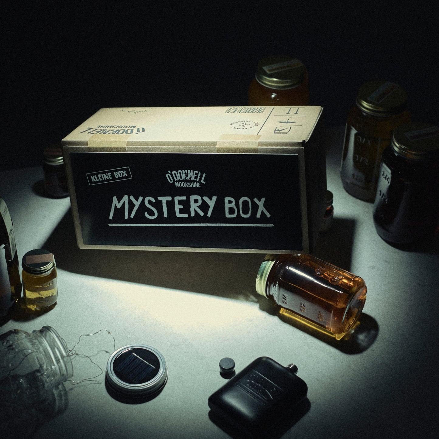Ein Lifestyle-Bild der kleinen Mystery Box. Die Mystery Box steht auf einem Tisch, umgeben von verschiedenen Gläsern und einigen Merchandise-Artikeln.