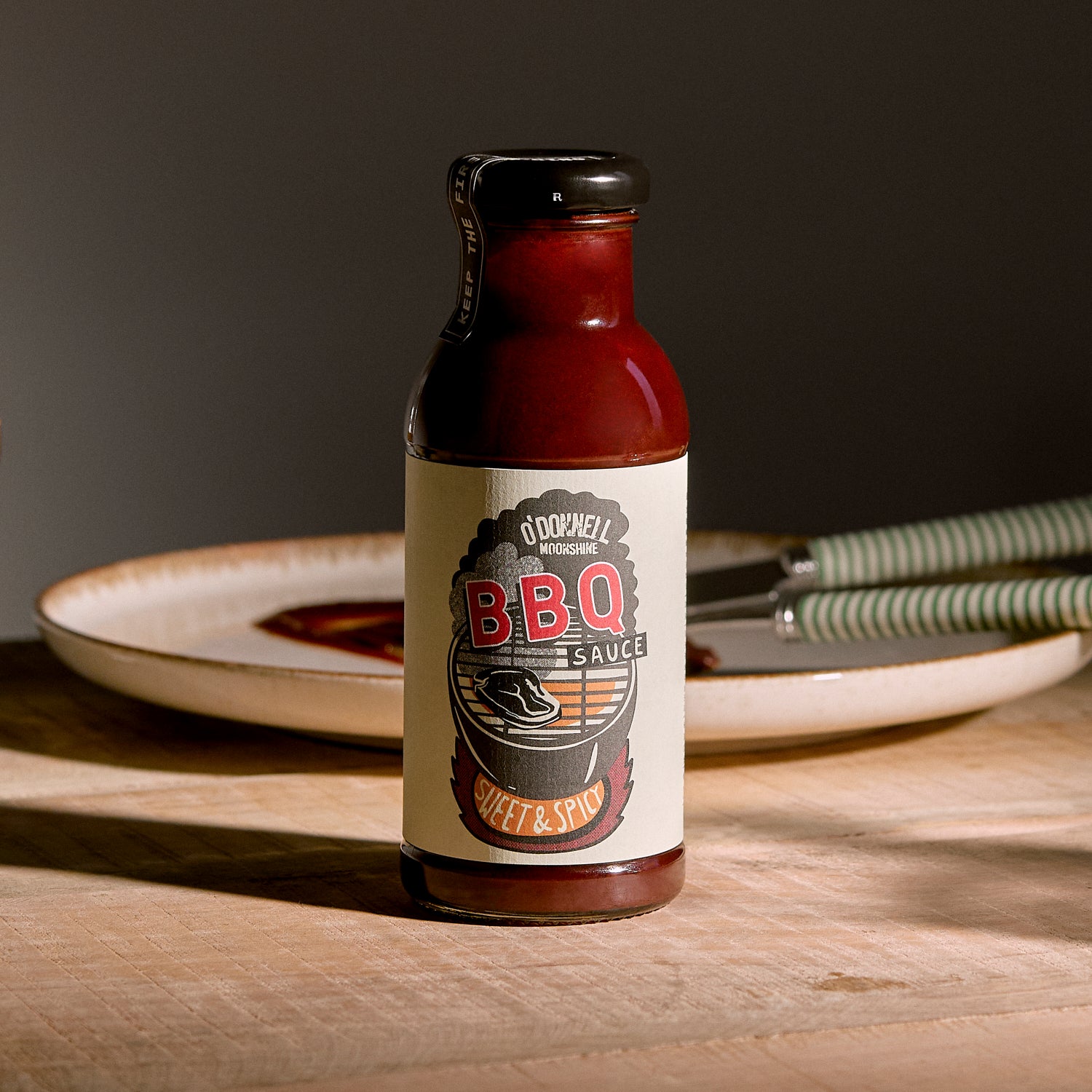 O'Donnell Moonshine BBQ Sauce Sweet & Spicy – Flasche auf Holzunterlage mit Tellern im Hintergrund, dunkle Studioatmosphäre