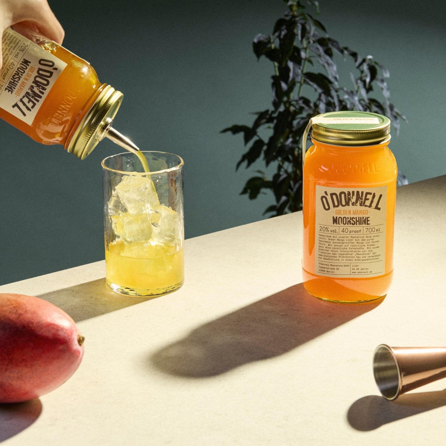 Auf der rechten Seite ein Glas Golden Mango Likör, auf der linken Seite ein Glas voller Eis und eine Person, die Mango Likör aus einem anderen Glas einschenkt.