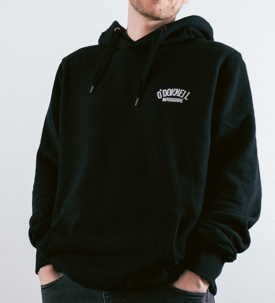 Mann trägt schwarzen O'Donnell Moonshine Hoodie – Frontalansicht mit weißem Logo-Aufdruck auf der Brust, helle Hintergrundaufnahme