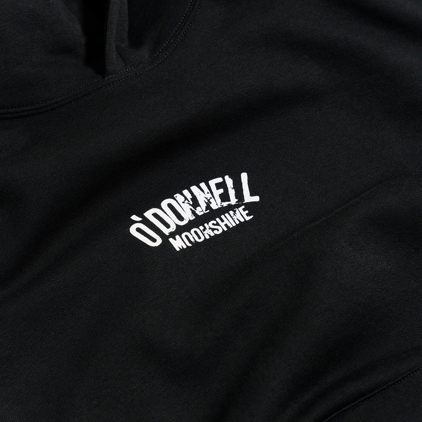 Quadratische Detailaufnahme für O'Donnell Moonshine: Ein weißer Logo-Print der Marke im Vintage-Stil prallt auf der Brust eines schwarzen Hoodies ab.