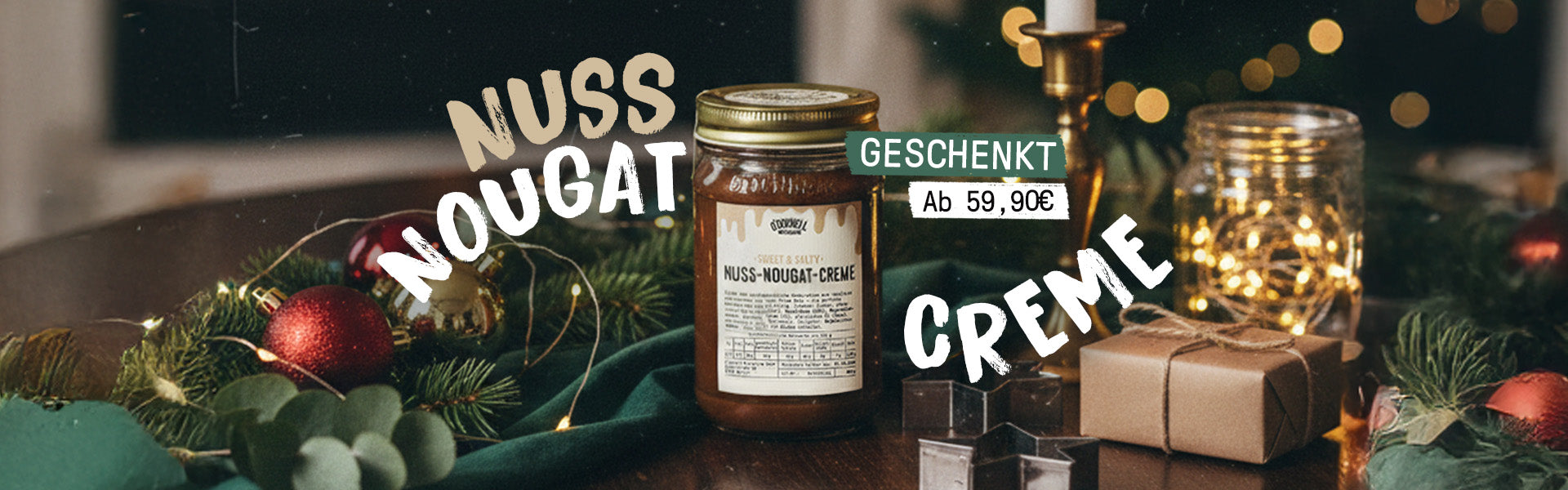 Breiter, dunkler und festlicher Weihnachts-Banner, der ein Glas Nuss-Nougat-Creme auf einem dunklen Holztisch zeigt. Er ist mit Tannenzweigen, roten Christbaumkugeln, Lichterketten und einem verpackten Geschenk dekoriert. Große Text-Overlays lauten „NUSS NOUGAT CREME“, und ein grünes Banner weist auf die Aktion hin: „GESCHENKT Ab 59,90 €“.