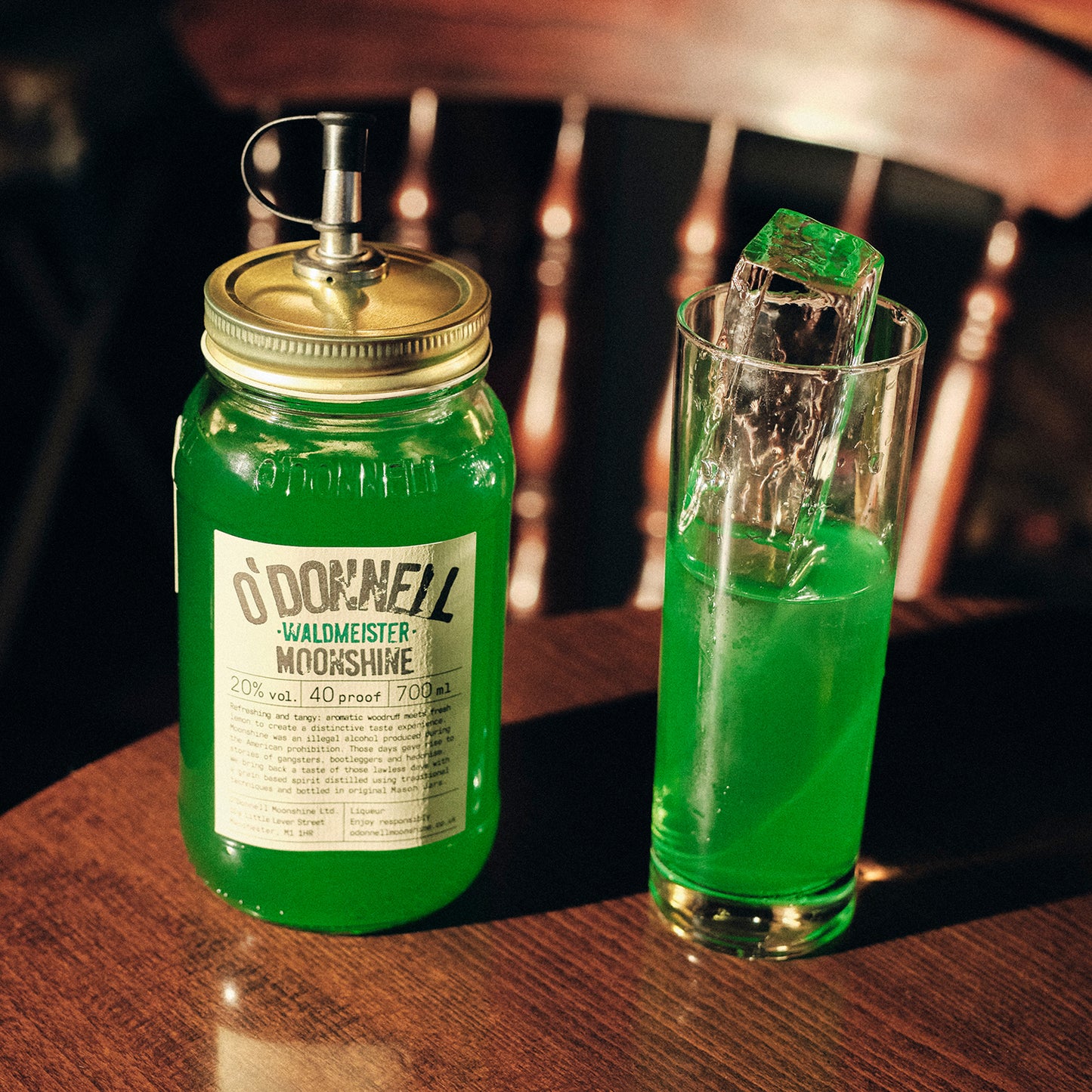 Ein großes, grünes Likörglas der Sorte „Waldmeister“ mit Ausgießer steht neben einem hohen Drink mit Eisstange auf Holz.