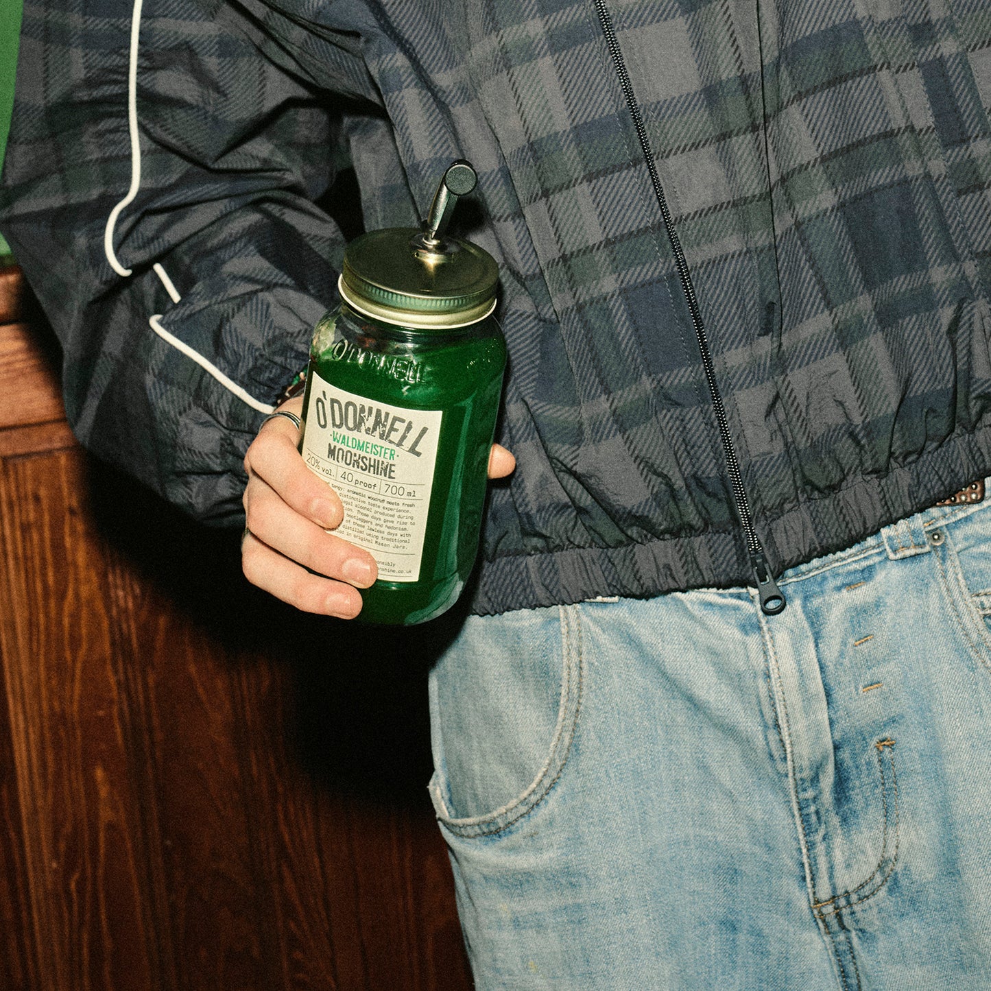 Nahaufnahme einer Person in karierter Jacke und Jeans, die ein großes, grünes O'Donnell „Waldmeister“ Likörglas mit Ausgießer in der Hand hält.