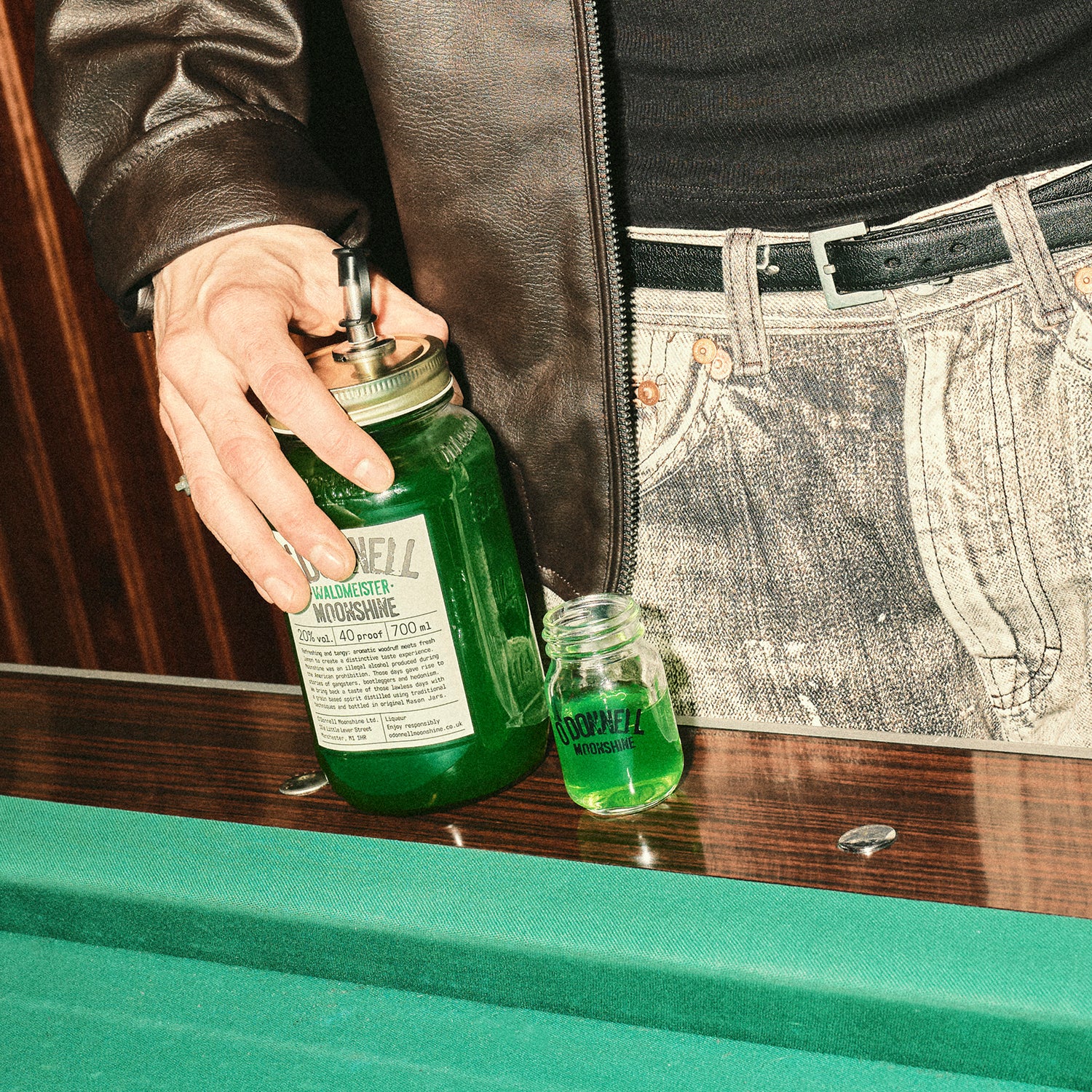 Nahaufnahme einer Hand, die ein großes grünes „Waldmeister“ Likörglas neben einem gefüllten Mini-Glas auf einer Billardtischkante hält.