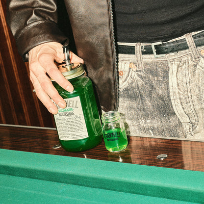 Nahaufnahme einer Hand, die ein großes grünes „Waldmeister“ Likörglas neben einem gefüllten Mini-Glas auf einer Billardtischkante hält.