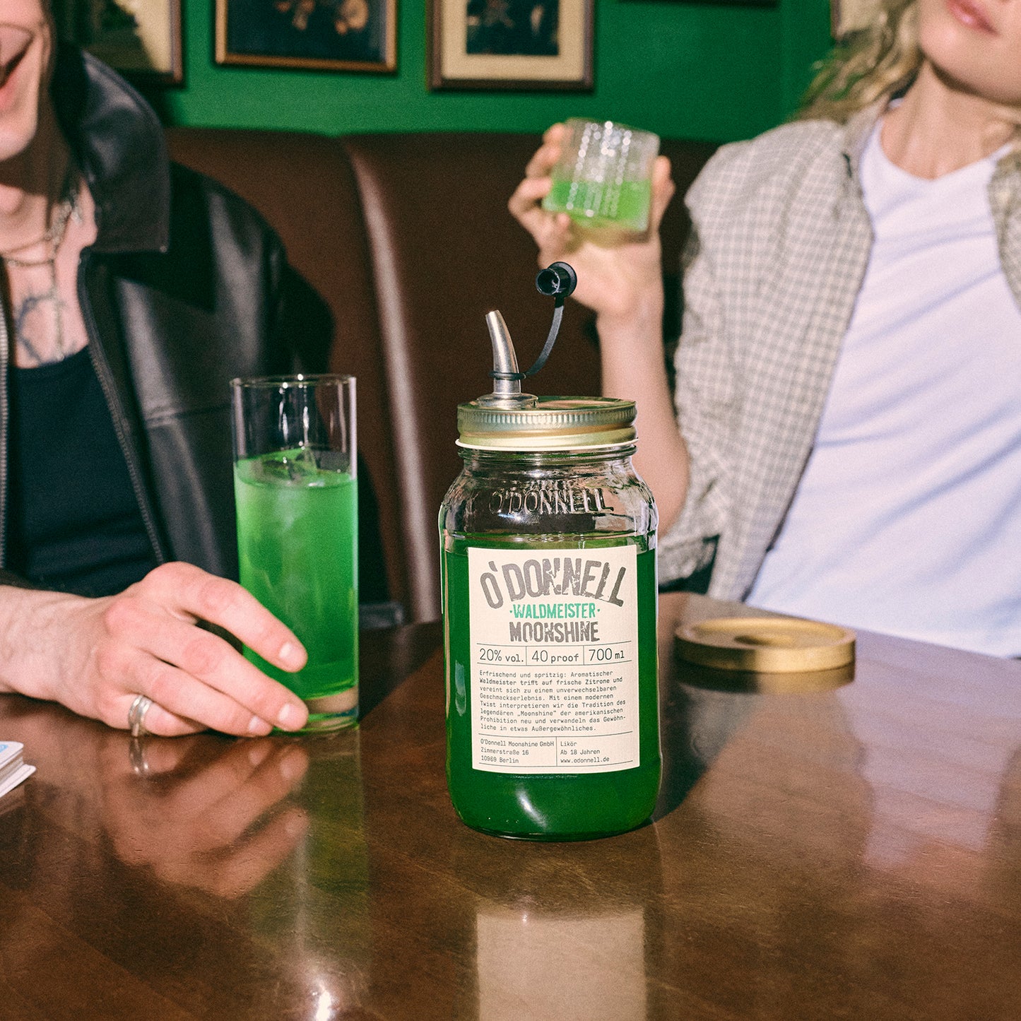 Ein großes O'Donnell „Waldmeister“ Likörglas steht im Fokus auf einem Tisch vor zwei Personen mit grünen Cocktails.