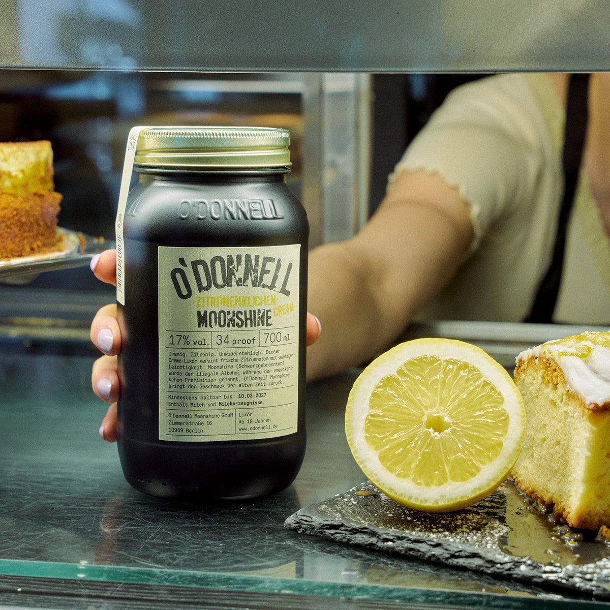 O'Donnell Moonshine Zitronenkuchen Cream 700ml in der Hand – Lifestyle-Produktbild in der Küche mit Zitrone und Zitronenkuchen
