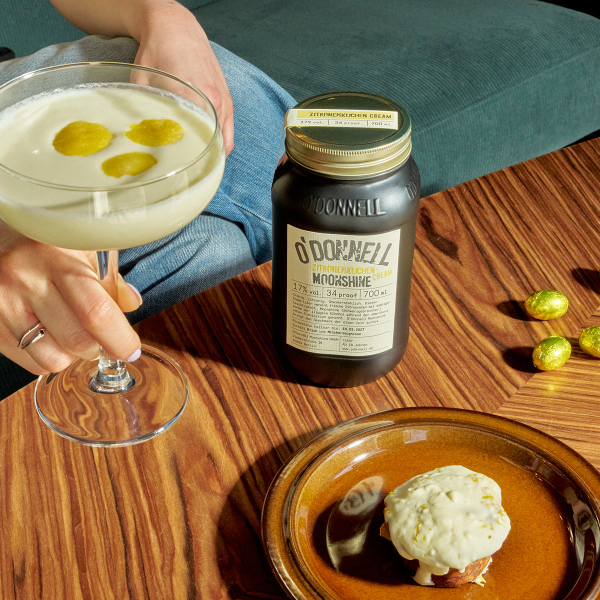 O'Donnell Moonshine Zitronenkuchen Cream 700ml – Lifestyle-Produktbild mit Cocktailglas, Zitronenkuchen und Osterschokolade auf Holztisch