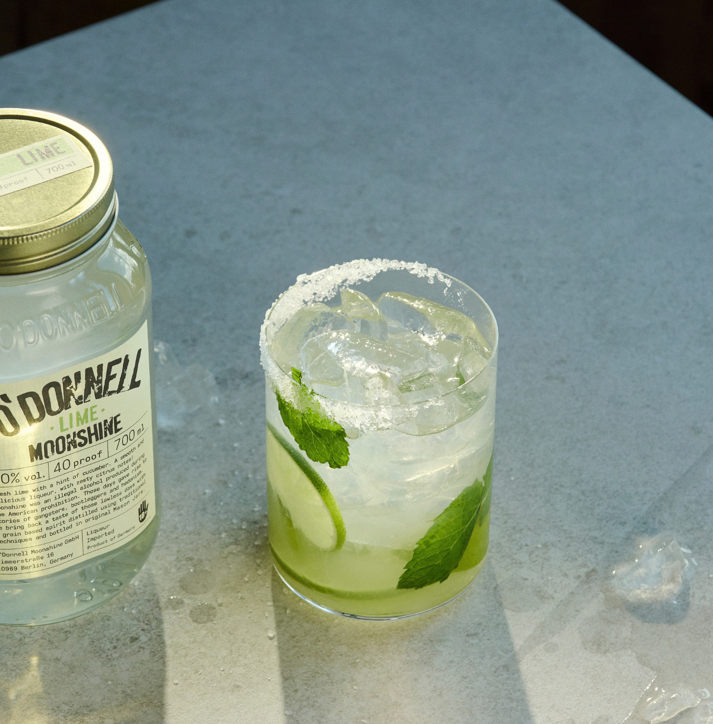 O'Donnell Moonshine Limette 700ml neben einem Cocktailglas mit Mojito-Stil, Minze, Limettenscheibe und Salzrand – Lifestyle-Foto auf Marmoroberfläche