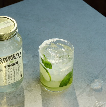 O'Donnell Moonshine Limette 700ml neben einem Cocktailglas mit Mojito-Stil, Minze, Limettenscheibe und Salzrand – Lifestyle-Foto auf Marmoroberfläche