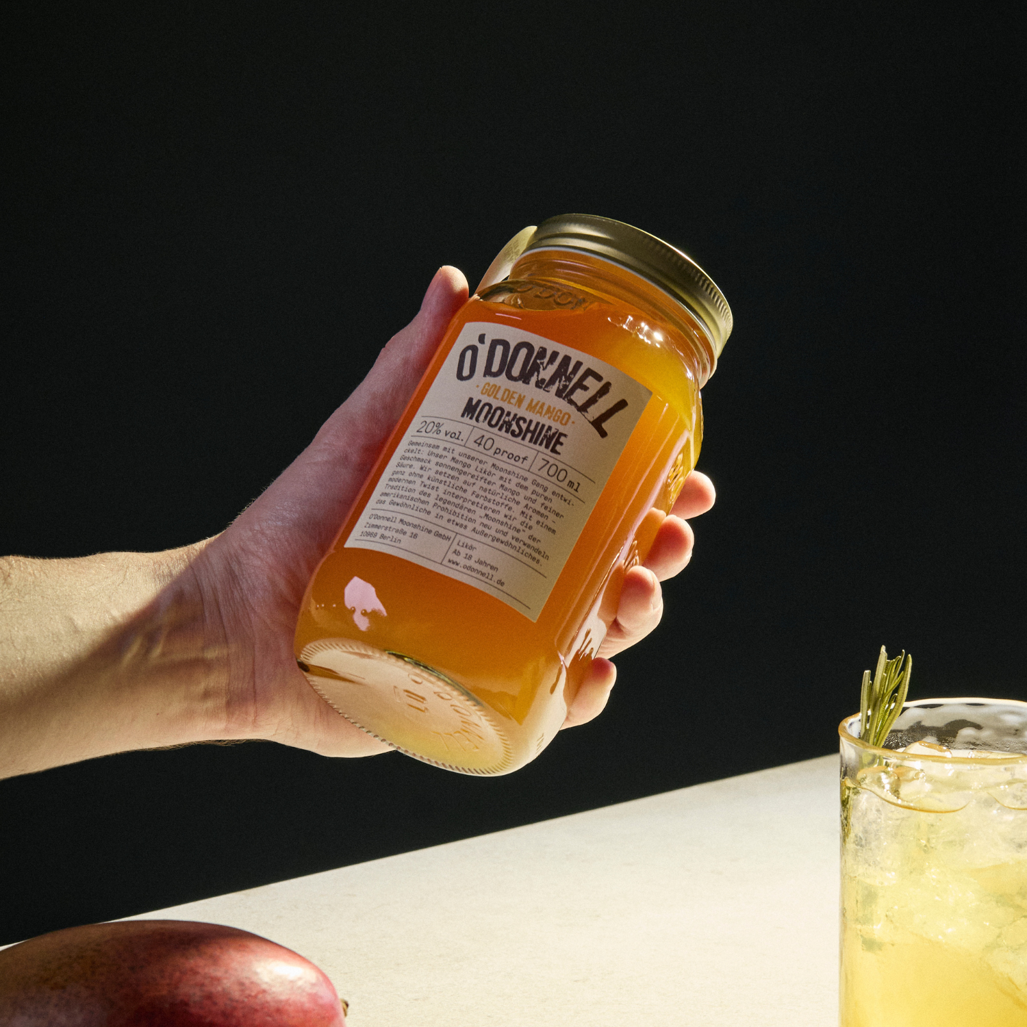 Eine Hand, die ein Glas Golden Mango Likör in warmen Licht hält.
