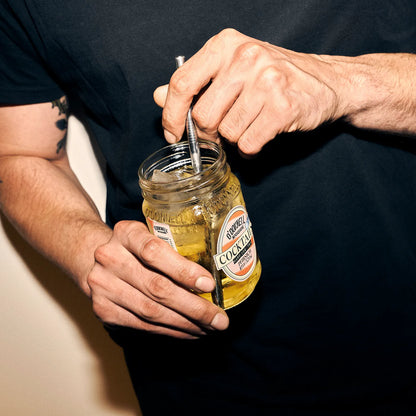 Quadratisches Lifestyle-Produktbild für eine Produktseite, das ein geöffnetes O'Donnell Moonshine Glas der Sorte „Cocktails Hard Ice Tea Pfirsich“ in den Händen einer Person zeigt. 