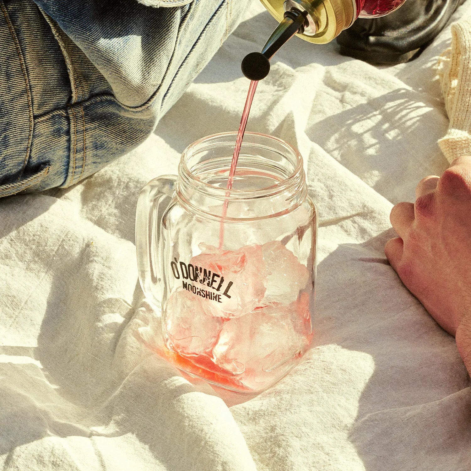 O'Donnell Moonshine Likeur wird in ein Henkelglas mit Eiswürfeln eingegossen – Outdoor-Lifestyle auf Leinentuch, Sonnenlicht