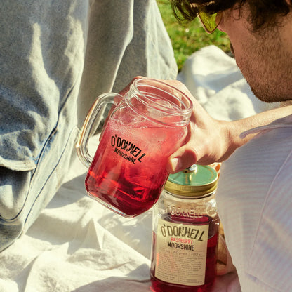 Mann hält O'Donnell Moonshine Henkelglas mit rotem Cocktail, daneben eine O'Donnell Moonshine Likeur-Flasche – Outdoor-Lifestyle, Draufsicht