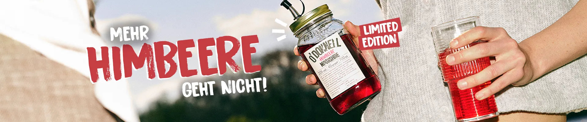 Eine Frau erhält O'Donnell Moonshine Himbeere Flasche und Cocktailglas im Freien vor blauem Himmel