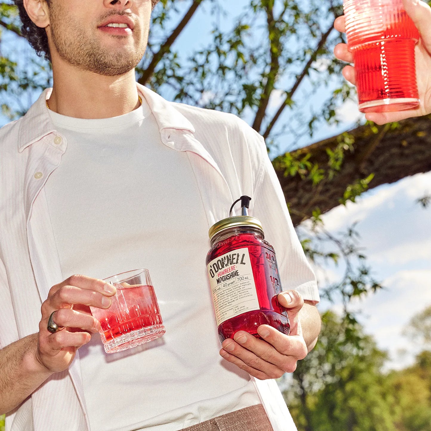 Mann hält O'Donnell Moonshine Himbeere Flasche und Cocktailglas im Freien unter Bäumen