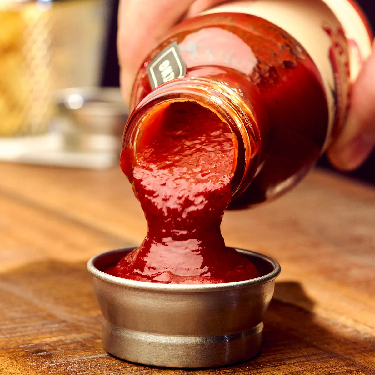 O'Donnell Moonshine BBQ Ketchup Pikant & Süss wird aus der Flasche in eine Metallschale gegossen – Nahaufnahme auf Holzuntergrund