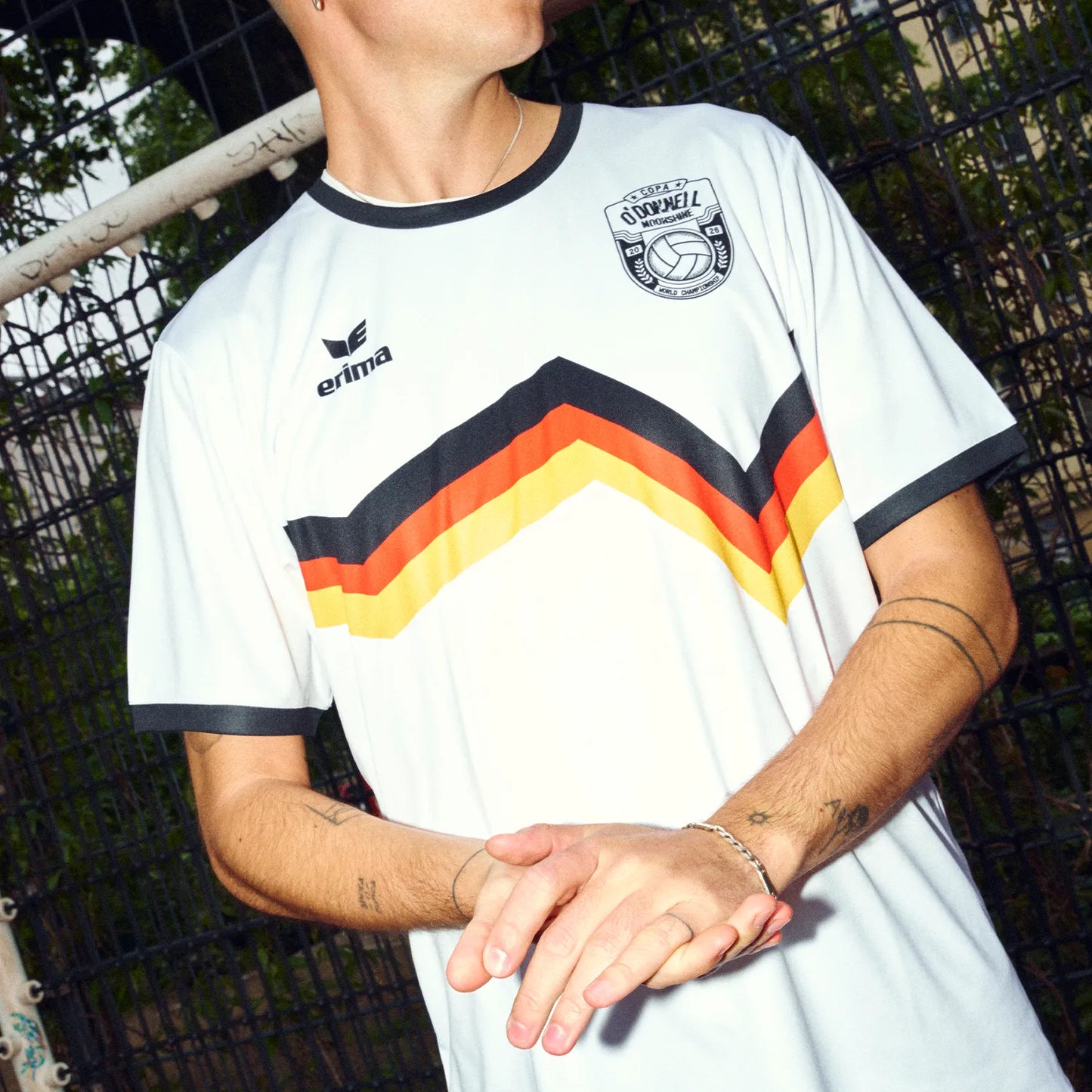 Mann trägt O'Donnell Moonshine x Erima WM Trikot DE – Nahaufnahme Torso mit schwarz-rot-gold Streifen und Copa-Badge, Outdoor vor Zaun