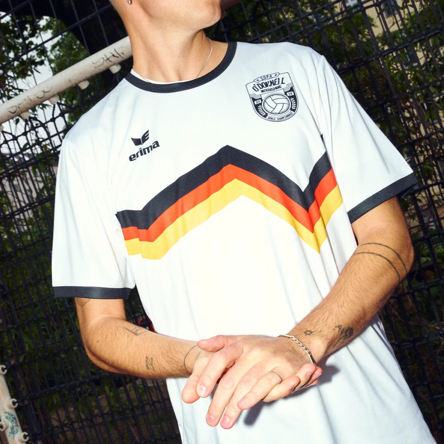 Mann trägt O'Donnell Moonshine x Erima WM Trikot DE – Nahaufnahme Torso mit schwarz-rot-gold Streifen und Copa-Badge, Outdoor vor Zaun