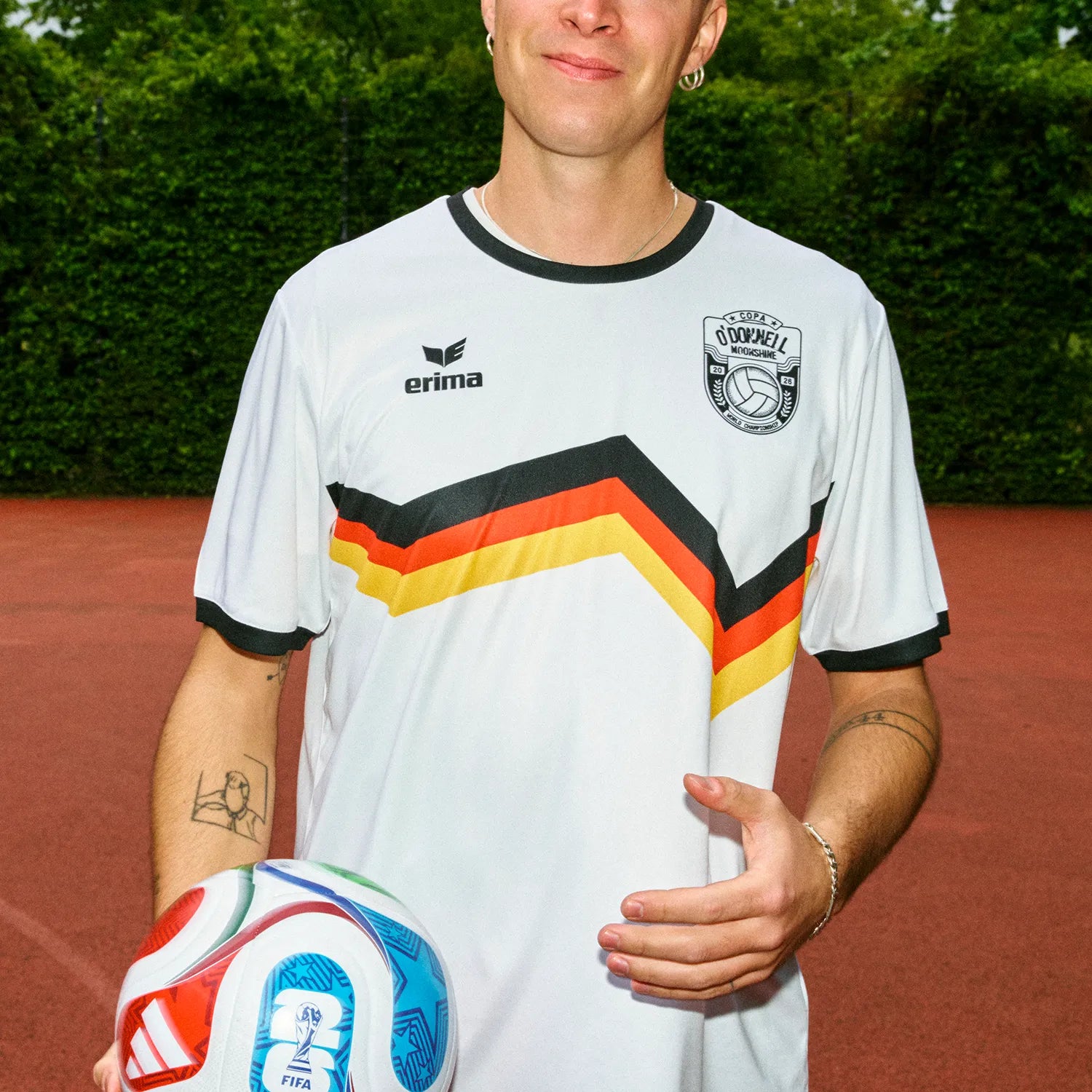 Mann trägt O'Donnell Moonshine x Erima WM Trikot DE – weißes Trikot mit schwarz-rot-gold Streifen, hält einen Fußball, Sportplatz im Hintergrund