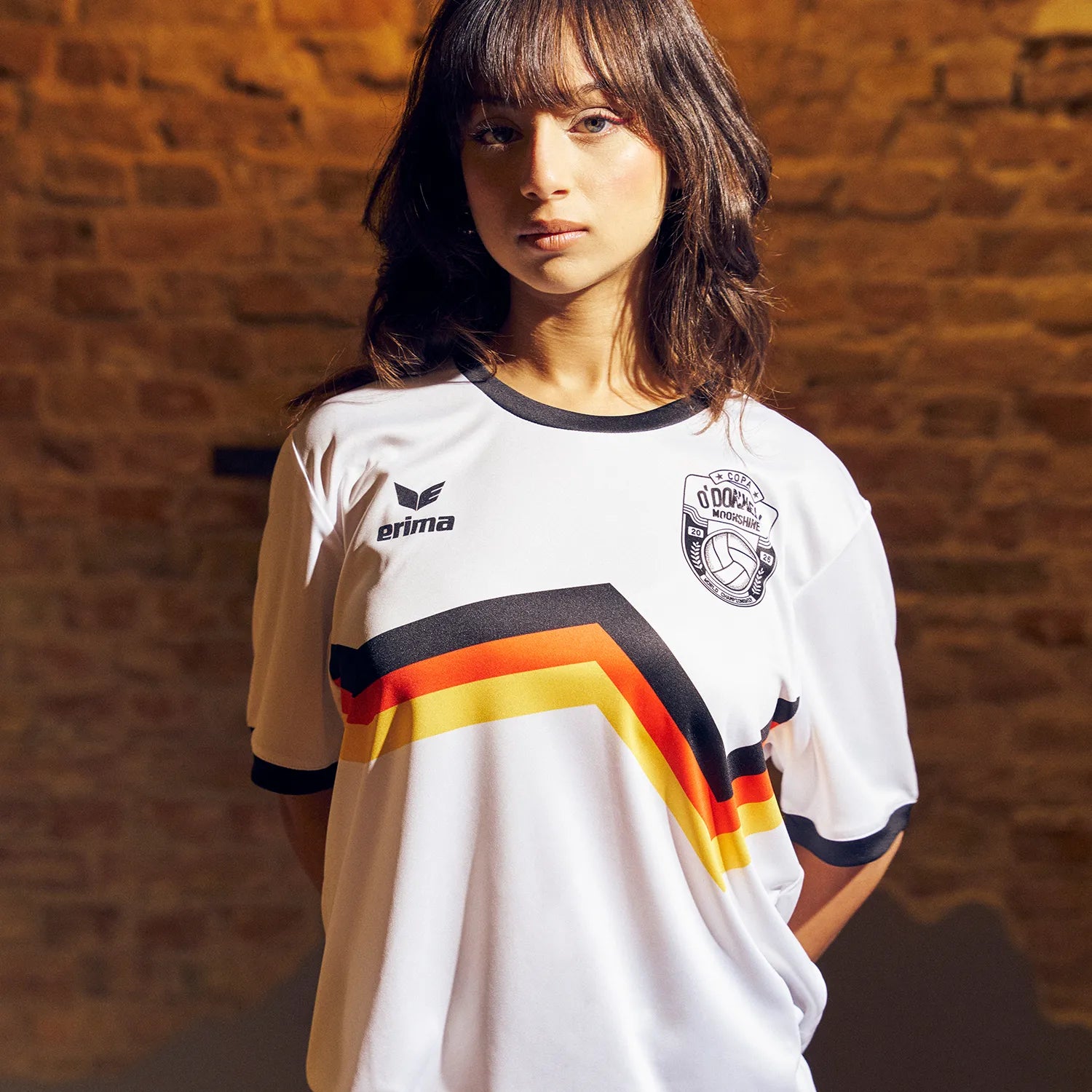 Frau trägt O'Donnell Moonshine x Erima WM Trikot – weißes Trikot mit deutschen Farben schwarz-rot-gold und Copa-Badge, vor Ziegelwand